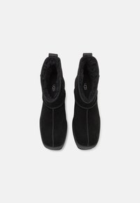 Bottines en daim noir avec doublure en peau de mouton, bout carré arrondi, languettes de tirage et logo UGG visible à l’intérieur de la semelle.