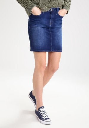 Person trägt einen grünen Strickpullover, einen blauen Jeansrock und dunkelblaue Canvas-Sneaker, steht mit den Händen in den Taschen.