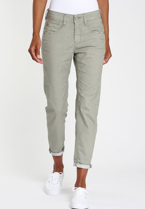 Femme portant un pantalon vert olive clair retroussé et des baskets blanches, debout devant un fond blanc uni.