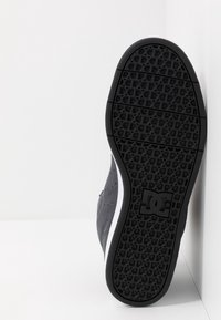 Musta kumipohja, jossa on geometrinen urituskuvio, valkoisella tehosteella sivussa ja tekstuurilla varustettu harmaa yläosa.