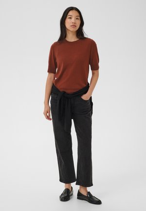 Femme portant un haut rouille à manches courtes, un pantalon noir à jambes larges avec un pull noir noué autour de la taille, et des mocassins noirs, se tenant devant un fond blanc.