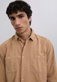 Beżowa koszula zapinana na guziki, wykonana z bawełny, z klasycznym kołnierzykiem, dwiema przednimi kieszeniami i luźnym krojem. Gładka tekstura i neutralny kolor.