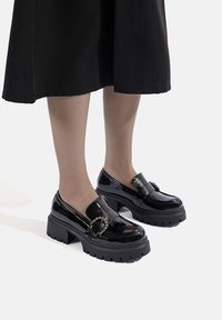 Mocassini in pelle verniciata nera con suola in gomma spessa, dotati di una fascia decorativa con piccoli dettagli e un design della punta arrotondato.
