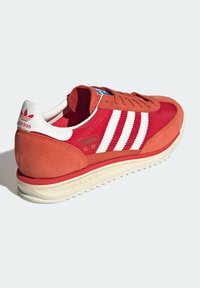 Röd mocka- och textilsko med vita tre ränder, vadderad krage och en kontrasterande benvit sula. Har "adidas"-logotyp på hälen.