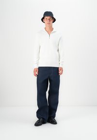 Suéter blanco de canalé con cuello de cremallera, combinado con vaqueros de pierna ancha azul oscuro y zapatos de cuero negros. El modelo lleva un sombrero bucket oscuro.