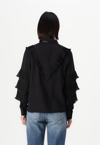 Camicia nera con maniche a pieghe voluminose e dettaglio a forma di V sul retro, realizzata in tessuto leggero, abbinata a jeans in denim azzurro chiaro.