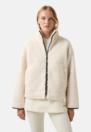 Blonde Frau, die eine cremefarbene Sherpa-Fleecejacke mit einem Reißverschluss vorne über einem hellen Hoodie und weißen Hosen trägt.