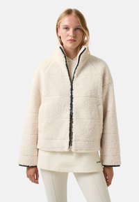 Blondynka w kremowej kurtce z owczej wełny z zamkiem błyskawicznym z przodu, nosząca jasny hoodie i białe spodnie.