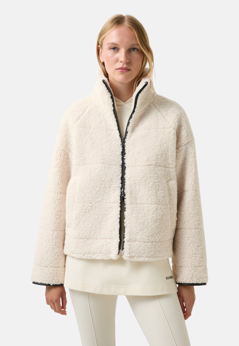 Blondynka w kremowej kurtce z owczej wełny z zamkiem błyskawicznym z przodu, nosząca jasny hoodie i białe spodnie.