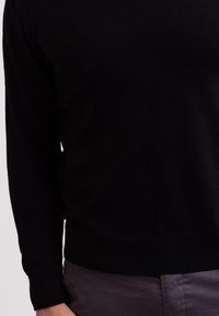 Schwarzer Pullover aus weichem Strickstoff, mit Rundhalsausschnitt und gerippten Bündchen und Saum. Kombiniert mit grauen Hosen.