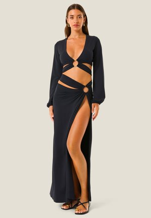 Donna che indossa un vestito nero a maniche lunghe con ritagli e dettagli ad anelli, con una spacco alto, abbinato a sandali neri con cinturini.