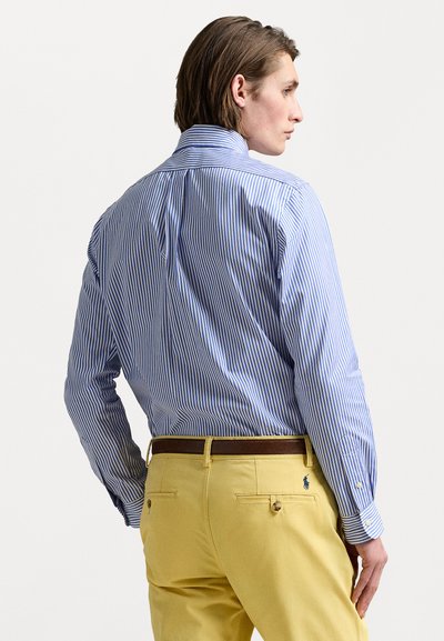 Prugačasta košulja na dugih rukava u plavoj i bijeloj boji, viđena s leđa, uparena s žutim hlačama i smešnim remenom. Prilagođeni kroj, dugi rukavi.