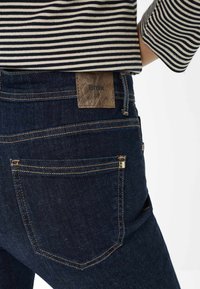 Jean en denim bleu foncé avec une patch en cuir marron portant l'inscrit "Brax", avec des coutures dorées et des détails de poche. Haut rayé visible.