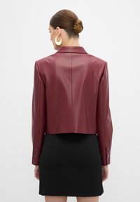 Blazer cropped in ecopelle bordeaux con colletto a revers, maniche lunghe e dettagli con bottoni, indossato sopra un vestito aderente nero.