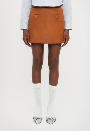 Brown suede mini skirt with two front pockets and round buttons, paired with white socks and silver pointy-toed shoes.