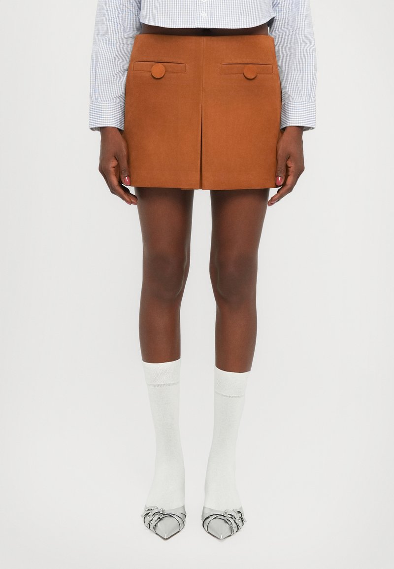 Mini gonna in suede marrone con due tasche frontali e bottoni rotondi, abbinata a calzini bianchi e scarpe argentate a punta.