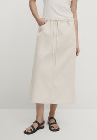 Beige, knælang nederdel lavet af bomuld. Har en elastisk talje med snøring og sidelommer. Glat tekstur.