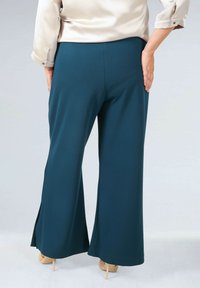 Teal wijde broek met een soepele textuur en subtiele zijsplitten, gecombineerd met een lichte zijden blouse. Schoenen zijn huidskleurige hakken.