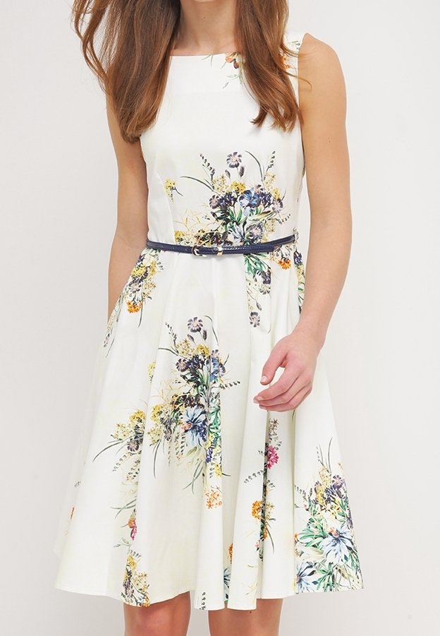 Femme portant une robe blanche sans manches avec des motifs floraux colorés et une fine ceinture sombre à la taille.
