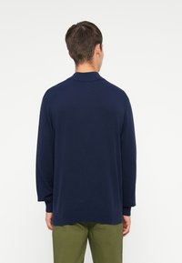 Granatowy sweter z dzianiny z wysokim kołnierzem, opadającymi ramionami i prążkowanymi mankietami. Materiał jest gładki, zapewniając luźny krój.