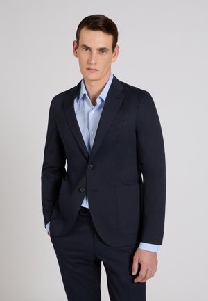 Giovane uomo che indossa un abito blu scuro con una camicia azzurro chiaro, in piedi davanti a uno sfondo grigio chiaro uniforme.