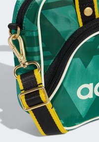 Πράσινη τσάντα με σχέδια και χρυσό κλιπ. Διαθέτει μαύρο ιμάντα με κίτρινες ρίγες και λευκή επένδυση κατά μήκος των άκρων. Ορατό το λογότυπο στο μπροστινό μέρος.