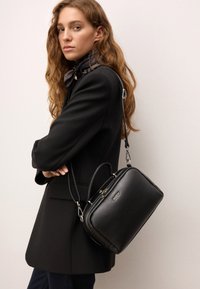 Črna usnjena torbica s strukturirano obliko, dvojni ročaji in odstranljiv ramenski trak. Ima srebrne kovinske dele in teksturirano površino.