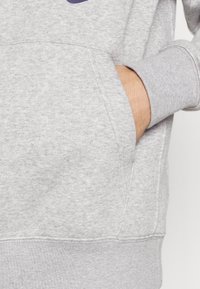 Main d'une personne partiellement dans la poche avant d'un sweat à capuche gris clair avec poignets et ceinture côtelés.