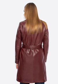 Donna con capelli lisci castano chiaro, che indossa un cappotto in pelle rosso scuro con cintura, in piedi di spalle contro uno sfondo bianco.