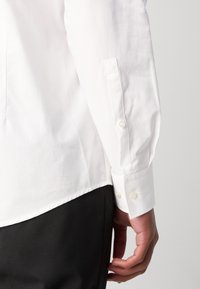 Gros plan sur le bras droit d'une personne portant une chemise blanche à manches longues avec poignet boutonné et un pantalon noir.