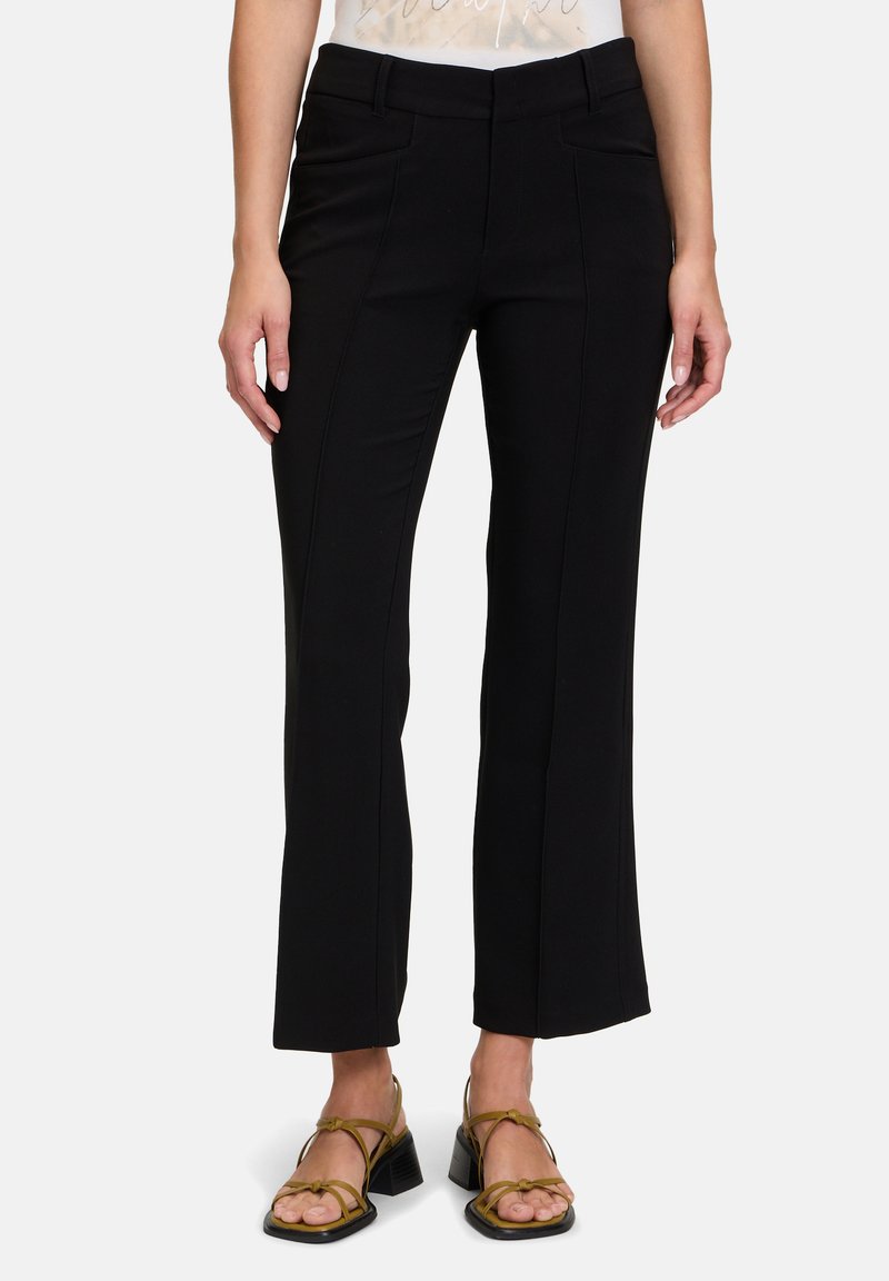 Schwarze maßgeschneiderte Hosen mit einem ausgestellten Bein-Design, ausgestattet mit Frontnähten und Taschen, aus einem glatten Stoff gefertigt. Getragen mit Sandalen mit Riemen.