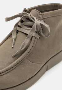 Gros plan sur une chaussure à lacets en daim beige avec des coutures épaisses et un embout métallique marqué Calvin Klein à une extrémité du lacet.