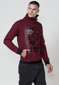 Sudadera roja con estampado y cuello alto, con detalles en negro. Presenta una impresión gráfica de calavera y texto. Combinada con jeans oscuros.
