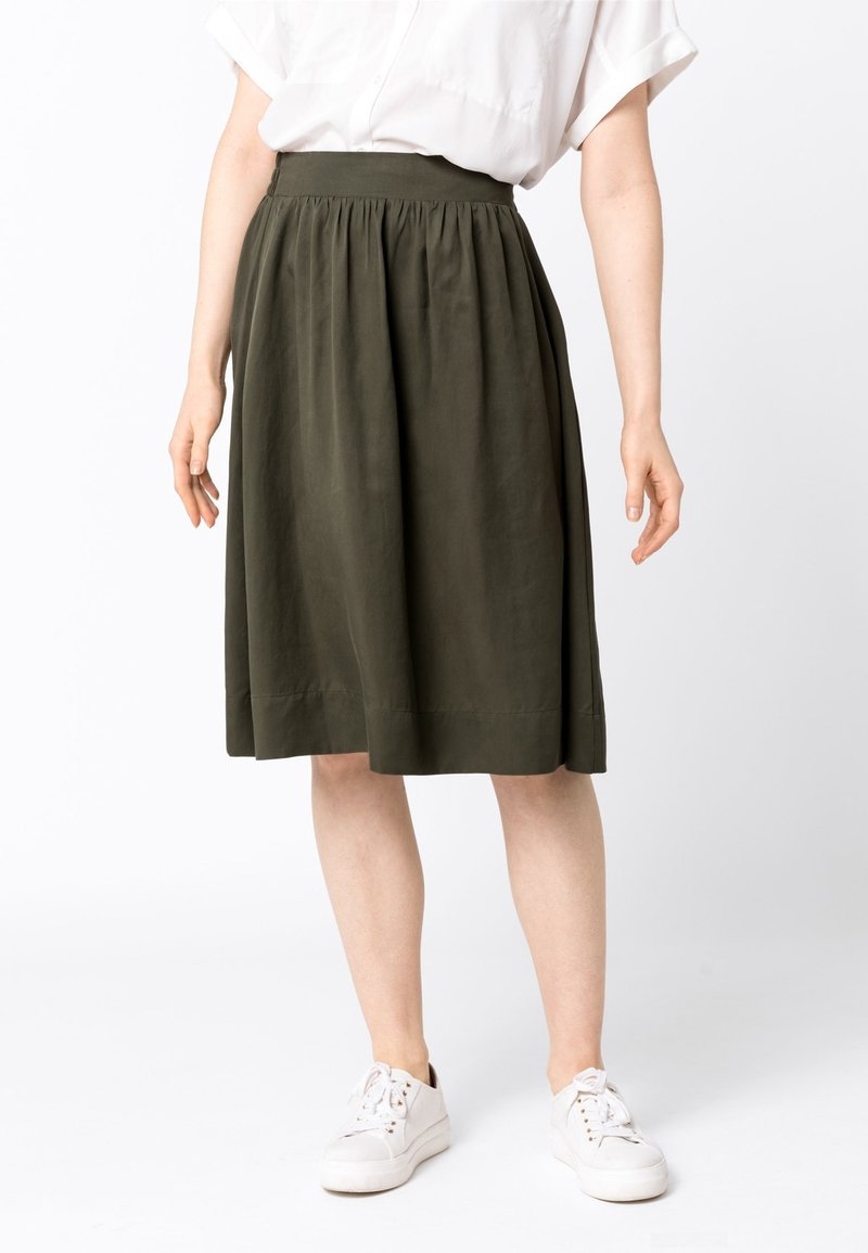 Femme portant une jupe longueur genou vert olive avec une taille froncée, un t-shirt blanc et des baskets blanches, debout devant un fond blanc uni.