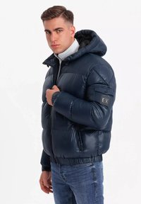 Giacca puffer blu navy con cappuccio, finitura lucida, chiusura con zip, maniche a polsino e dettaglio con patch sul braccio sinistro. Indossata sopra una maglietta grigia chiara.