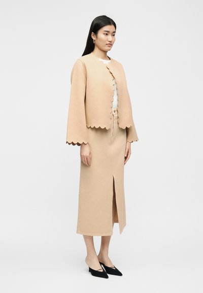 Vrouw staand, gekleed in een beige jas met een gegolfde rand en een bijpassende midi-rok met een split aan de voorkant, gecombineerd met zwarte puntige hakken.