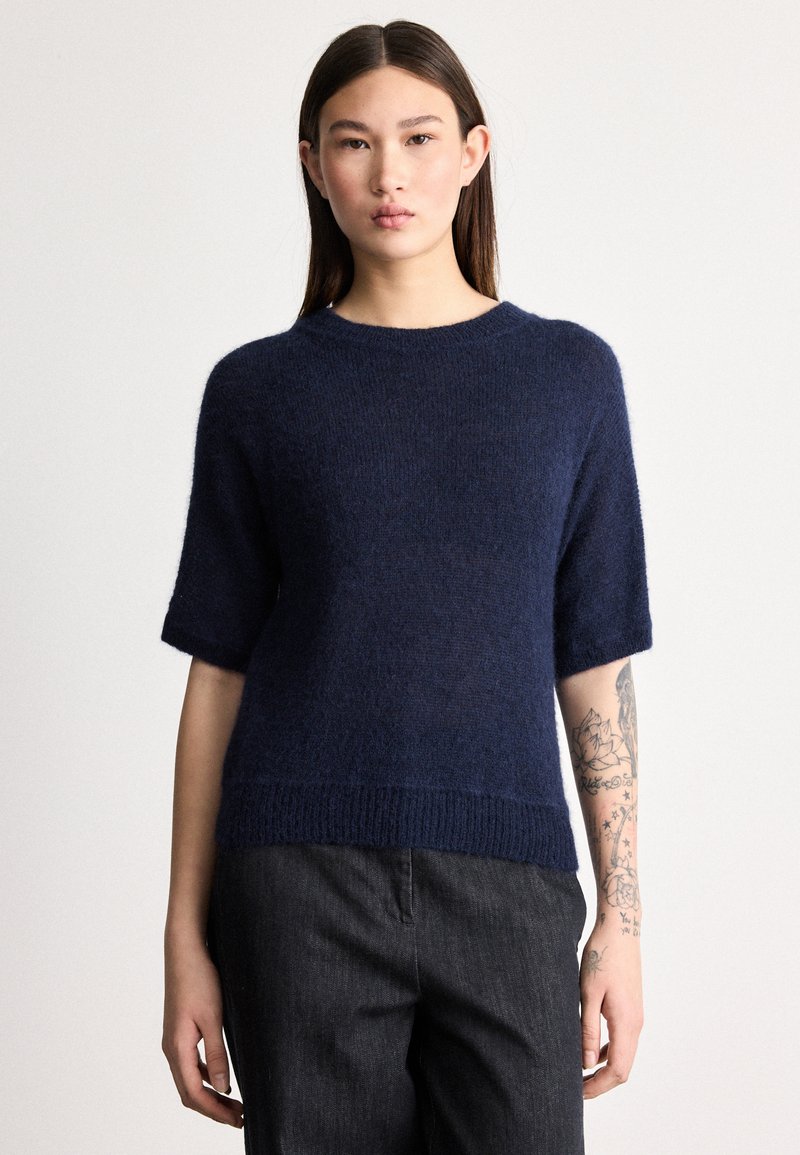 Πλεκτό πουλόβερ σε ναυτικό μπλε χρώμα με κοντά μανίκια, στρογγυλή λαιμόκοψη και ριμπ τελείωμα. Απαλή υφή με χαλαρή εφαρμογή. Το μοντέλο φοράει σκούρο γκρι παντελόνι.