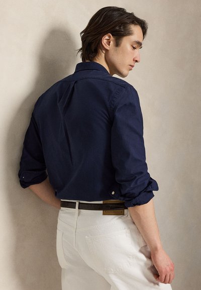 Jaunas vyras iš šono, dėvintis susuką tamsiai mėlyną marškinių rankovę, įkištą į baltas kelnes su rudais diržu priešais smėlio spalvos sieną.