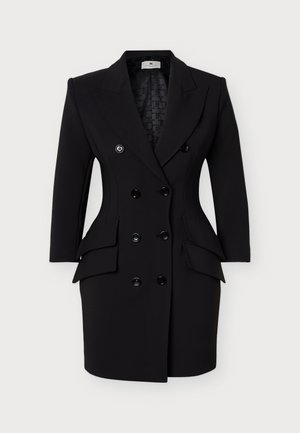 Femme portant une robe blazer noire à double boutonnage avec poches à rabat et des bottes noires montantes à talons, debout devant un fond uni.