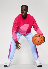Felroze fleece sweatshirt met een groot roze patroon, gecombineerd met lichtblauwe joggingbroek met paarse zijstripes. Houdt een basketbal vast.