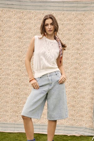 Tânără purtând un top alb fără mâneci tricotat, pantaloni scurți din denim deschis până la genunchi și un fular cu model în păr, stând în fața unui fundal floral.