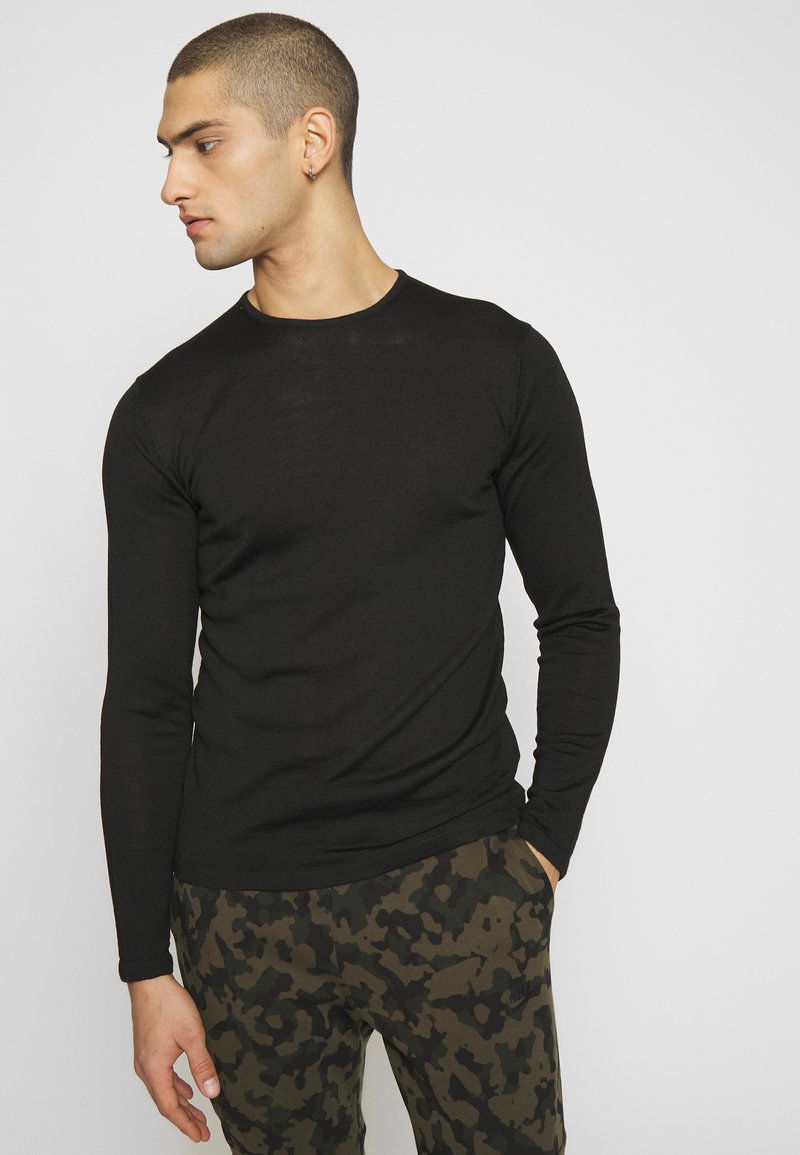 Chemise noire à manches longues avec col rond, associée à un pantalon camouflé vert avec des accents noirs et un logo. Texture lisse sur l'ensemble.