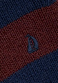 Strickstoff in marineblauen und dunkelroten Streifen. Verfügt über ein kleines, gesticktes Segelboot-Logo in dunklerem Blau. Die Textur wirkt weich und dick.