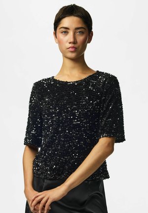 Femme aux cheveux courts et foncés portant un haut noir à manches courtes pailleté et un pantalon noir, debout les mains jointes devant.