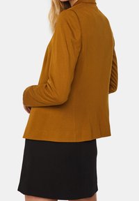 Hořčicově žlutý blazer s dlouhými rukávy, hladkou texturou a přiléhavým střihem, spárovaný s černou sukní, z pohledu zezadu.