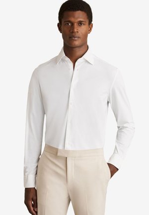 Homme portant une chemise blanche à manches longues boutonnée, rentrée dans un pantalon beige, debout avec une main dans la poche.