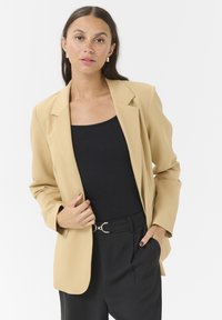 Blazer beige con spalle strutturate e una vestibilità rilassata. Abbinato a una maglia nera e pantaloni neri a vita alta, con una fibbia argentata.