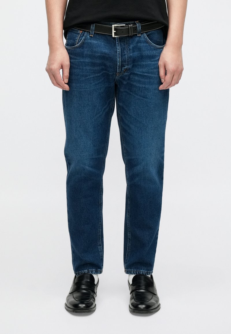 Jean droit en denim bleu moyen, avec cinq poches, une braguette zippée et un léger délavage. Porté avec une ceinture noire et des chaussures.