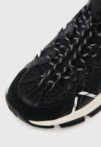 Black athletic shoe with mixed textures, featuring mesh and suede, white accents, patterned laces, and a contoured rubber sole.