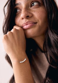Silver bracelet with five white pearls, delicate chain design; worn on a light-skinned wrist. Background is blurred neutral tones.